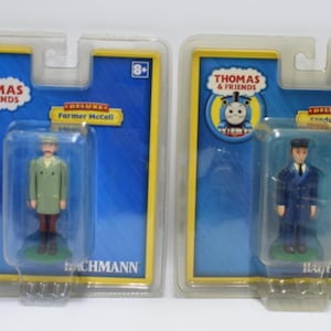 May include: Two Deluxe Thomas & Friends figurines in packaging. One is Farmer McColl in a green coat and brown pants. The other is a Conductor in a blue uniform. Both are in clear plastic packaging with the Thomas & Friends logo.