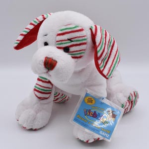 Peut inclure: Un jouet en peluche en forme de chien blanc avec des accents rayés rouges et verts sur les oreilles, les yeux, le nez et les pattes. Le jouet porte une étiquette "Webkinz".