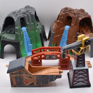 May include: A toy train set with mountain tunnels, a bridge, and a crane. The mountain tunnels are gray and brown with blue waterfalls. The bridge is orange with the text "VICKSTOWN DIESELWORKS". The crane is yellow and gray.
