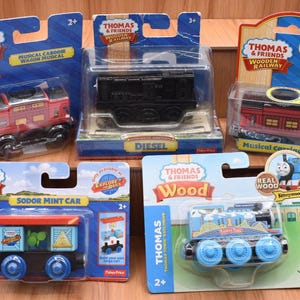 May include: A collection of Thomas & Friends wooden railway toys, including a red musical caboose, a black Diesel engine, and a blue and red Thomas the Tank Engine. Each toy is packaged in a clear plastic case.