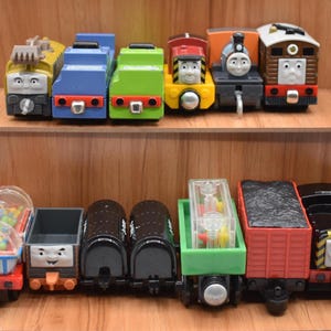 Peut inclure: Collection de locomotives et de wagons de train jouets colorés, dont Diesel, Thomas et Toby. Les trains sont disposés sur des étagères en bois, présentant divers modèles et personnages d'une série pour enfants.