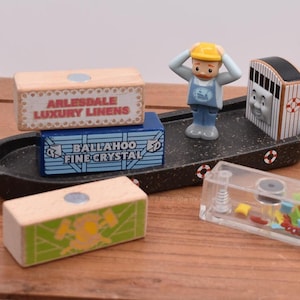 Może przedstawiać: Drewniana zabawka w kształcie łodzi z figurką mężczyzny w żółtym kasku i wagonem z kreskówki. Znajdują się trzy drewniane pudełka z napisem: "ARLESDALE LUXURY LINENS" i "BALLAHOO FINE CRYSTAL". Obecny jest również przezroczysty plastikowy pojemnik.