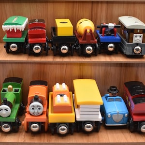 Può includere: Una collezione di colorati trenini giocattolo in legno esposti su scaffali di legno. I treni presentano vari design, tra cui una locomotiva verde, un vagone rosso e un'auto blu con un conducente. Ogni treno ha una forma e un colore unici.