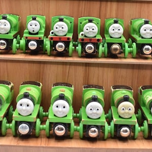 May include: A collection of twelve green toy train engines featuring white faces with various expressions. Each engine has a cylindrical body, a black chimney, and four silver-coloured wheels. The trains are arranged in two rows of six on wooden shelves.