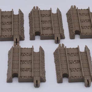 May include: Five brown plastic toy train track pieces. Each piece has a rectangular shape with parallel tracks and a textured, stone-like border. The tracks are designed to connect to each other.