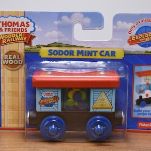 May include: A Thomas & Friends wooden railway toy train car, the "Sodor Mint Car", packaged in a blue and red cardboard. The car is light blue with a red roof and blue wheels. The packaging includes the text "Explore the Rails".