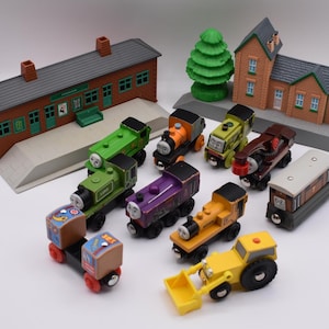 May include: A selection of wooden toy trains, a yellow tractor, and model railway buildings. The trains are in various colours, including green, purple, orange, and yellow. The buildings are brick-coloured with grey roofs and green accents.