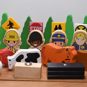 May include: A collection of wooden toy figures and accessories, including people, animals, road signs, and a fuel tank. The figures depict various professions, such as a police officer and a firefighter. The toys are arranged on a wooden surface.