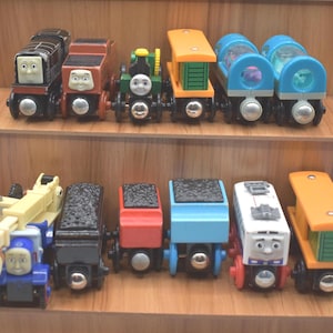 May include: A collection of colorful wooden toy trains, including engines and cars, displayed on wooden shelves. The trains feature various colors like blue, red, green, and orange, with detailed faces and designs.