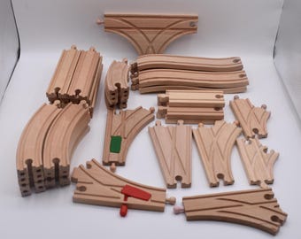 May include: A collection of light-colored wooden toy train track pieces, including straight segments, curved sections, Y-shaped switches, and a T-junction. One switch features a red plastic lever, and another includes a small green plastic insert.