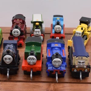 May include: A selection of toy train engines in various colours, including red, blue, green, and yellow. The engines are arranged on a wooden surface, showcasing their detailed designs and friendly faces. A green passenger carriage is also present.