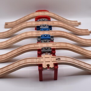 May include: A set of five arched wooden train track segments arranged in parallel, each supported by a plastic bridge pier. The piers are coloured red, blue, and grey, featuring a central cross-track piece at the base of the arrangement.