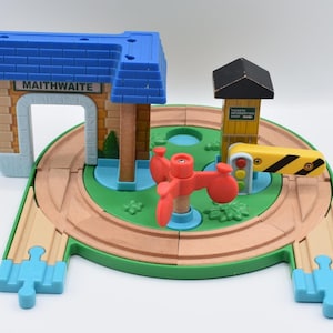 May include: A wooden toy train set featuring a station with "MAITHWAITE" text, a signal, and a crossing gate. The set includes a circular track, a red windmill, and a blue roof. The track is surrounded by green and blue elements.