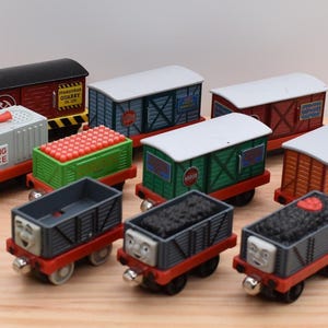 May include: A collection of nine toy train carriages featuring various designs, including a grey warning device carriage with red text, a green carriage filled with red cargo, and several freight carriages in red, blue, green, and orange with painted faces and coal loads.