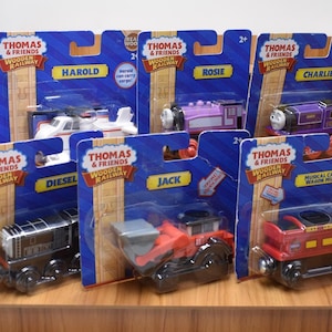 Może przedstawiać: Asortyment zabawek Thomas & Friends Wooden Railway, w tym Harold helikopter, Rosie fioletowy pociąg i Diesel czarny pociąg. Każda zabawka jest zapakowana na niebieskim kartonowym podkładzie z imieniem postaci.