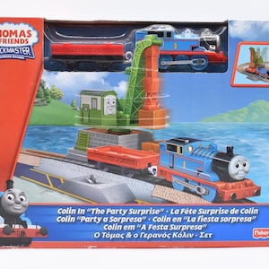 Könnte beinhalten: Ein Thomas & Friends Trackmaster Spielzeugset mit einer blauen Thomas die Tank Engine Lokomotive, einem grünen Kran und roten Güterwagen. Auf der Box steht "Colin In 'The Party Surprise'" und ein Bild von Thomas.