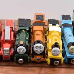 May include: A selection of seven brightly coloured wooden toy trains, each with a smiling face. The trains are red, orange, green, yellow, and blue, arranged on a wooden surface.