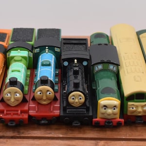 May include: A selection of toy train engines in various colours, including orange, green, blue, black, and yellow. Each train has a distinctive face. The toys are arranged on a wooden surface.