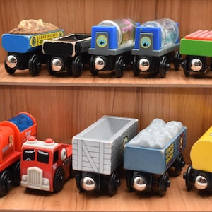 May include: A selection of brightly coloured wooden toy train carriages. The top row includes a blue carriage with a brown load, a black carriage, two blue carriages with clear tops, and a green carriage with a red top. The bottom row features a red fire engine, a red fire truck, a grey boxcar, a blue coal car, and a yellow tanker car.
