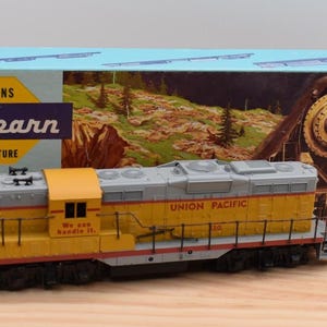 May include: A Union Pacific model diesel locomotive in yellow and grey with black railings and wheels. The side of the cab features the text We can handle it. The train is positioned in front of its original light blue Athearn Trains in Miniature box.