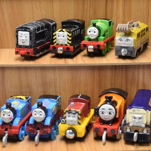 Peut inclure: Plusieurs modèles de trains jouets aux couleurs variées, dont le noir, le vert, le bleu et le jaune. Chaque locomotive présente un visage et un design uniques, avec des détails tels que des roues rouges et des finitions argentées. Les trains sont disposés sur des étagères en bois.