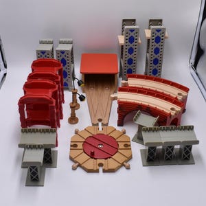 May include: A selection of wooden toy train track components, including bridges, a station, and supports. The set features red, tan, and grey pieces, with a circular track piece and a small lamp post. The pieces are designed for a toy train set.