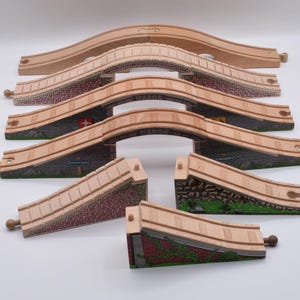 May include: A collection of wooden toy train track pieces including arched bridge sections and sloped ramp supports. The bridges feature printed brick and stone patterns, with text labels reading Maron, Knapford, and Crosby visible on the sides.
