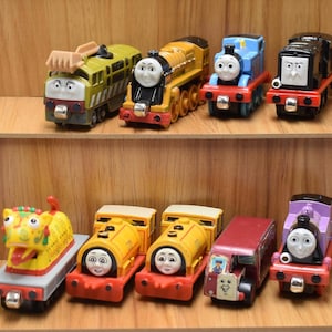 May include: A selection of toy train engines in various colours, including blue, yellow, green, and purple. The engines have smiling faces and are arranged on wooden shelves. The toys are designed for children.