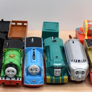 Könnte beinhalten: Eine Sammlung von sechs Spielzeugeisenbahnen und Waggons, darunter eine schwarze Dampflokomotive, eine grüne, eine blaue, eine türkisfarbene, eine silberne stromlinienförmige Lokomotive und eine beige mit Partyhut, alle mit Gesichtern.