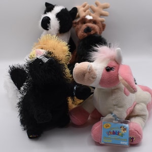Puede incluir: Una colección de animales de peluche, que incluye una vaca negra y blanca, un reno marrón, una mofeta negra y blanca, un gato amarillo y un caballo rosa y blanco. El caballo tiene una etiqueta que dice "Webkinz".