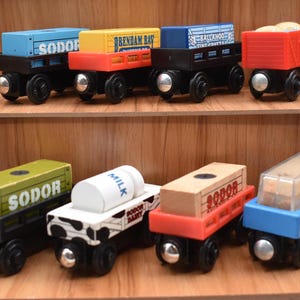 May include: A selection of brightly coloured wooden toy train carriages. Each carriage has a different coloured top and is labelled with various names. The carriages are on a wooden shelf, a classic toy.