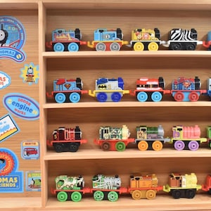 May include: A wooden display case filled with colorful toy train engines. The trains are arranged on shelves and feature various designs and characters. Stickers with the Thomas & Friends logo are on the side.