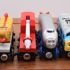 May include: A collection of six colorful wooden toy trains. Each train features a unique design, including a yellow and red dragon, a red and white train, and a silver train with a face. The trains are on a wooden surface.