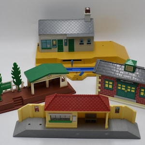 May include: A collection of toy train station buildings in various styles. The set includes a station with a red roof, a brick building with a green door, and a building with a green roof. The buildings are made of plastic.