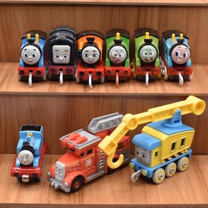 May include: A collection of nine toy trains and vehicles featuring various characters with distinct facial expressions. The set includes six small locomotives in blue, black, orange, green, and light blue, plus a blue engine, a red fire engine, and a yellow crane truck.