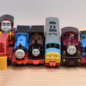 May include: A collection of eight toy trains featuring various characters with painted faces. The set includes blue, red, brown, grey, and purple engines with black wheels and red chassis, arranged in a row on a light-coloured wooden surface.