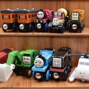 May include: A collection of colorful wooden toy trains and vehicles, including a red train, a blue train, a green excavator, and a white helicopter. The toys are arranged on wooden shelves.