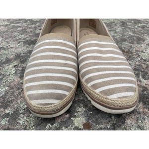 May include: Pair of beige and white striped slip-on shoes. The shoes have a rounded toe, a white sole, and a woven jute trim. The upper part of the shoe features horizontal stripes in beige and white.