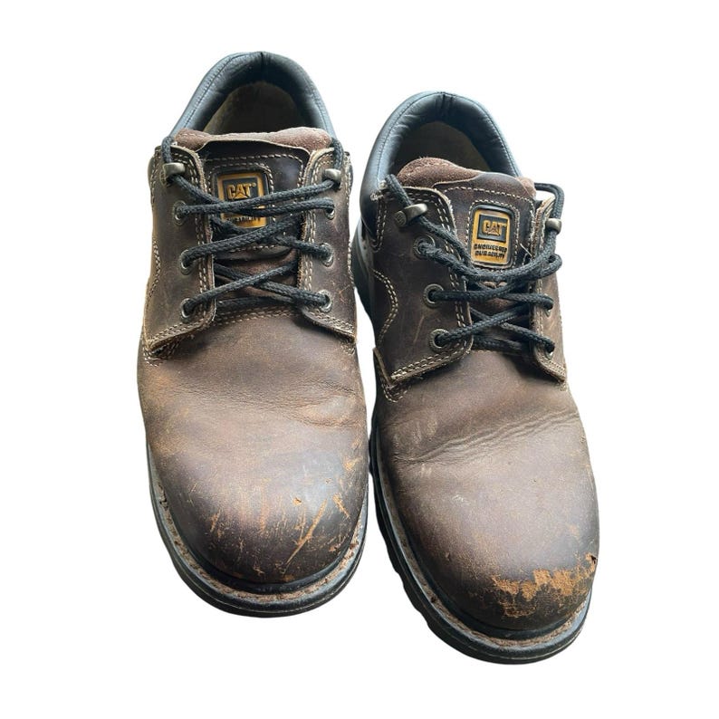 May include: Pair of brown leather work shoes with black laces. The shoes show signs of wear, with scuffs and wear visible on the leather. The Caterpillar logo is visible on the tongue of each shoe. The soles are black and thick.