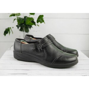 May include: A pair of black leather slip-on shoes with a low heel. The shoes have a textured surface and a decorative strap with a buckle. The shoes are displayed on a white wooden surface.