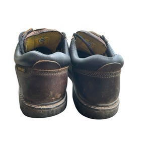 May include: Pair of brown leather work boots with black trim. The boots have a worn appearance and feature the Caterpillar logo on the side. The interior is yellow and the laces are black.