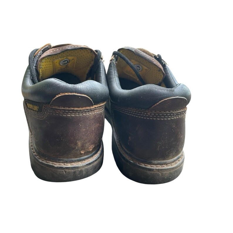 May include: Pair of brown leather work boots with black trim. The boots have a worn appearance and feature the Caterpillar logo on the side. The interior is yellow and the laces are black.