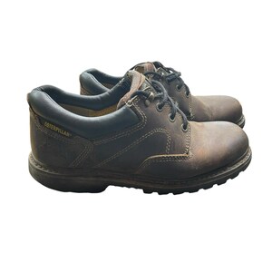 May include: Pair of brown leather work boots with black laces. The boots have a rounded toe and a thick, dark brown sole. The brand name "Caterpillar" is visible on the side of the boot.