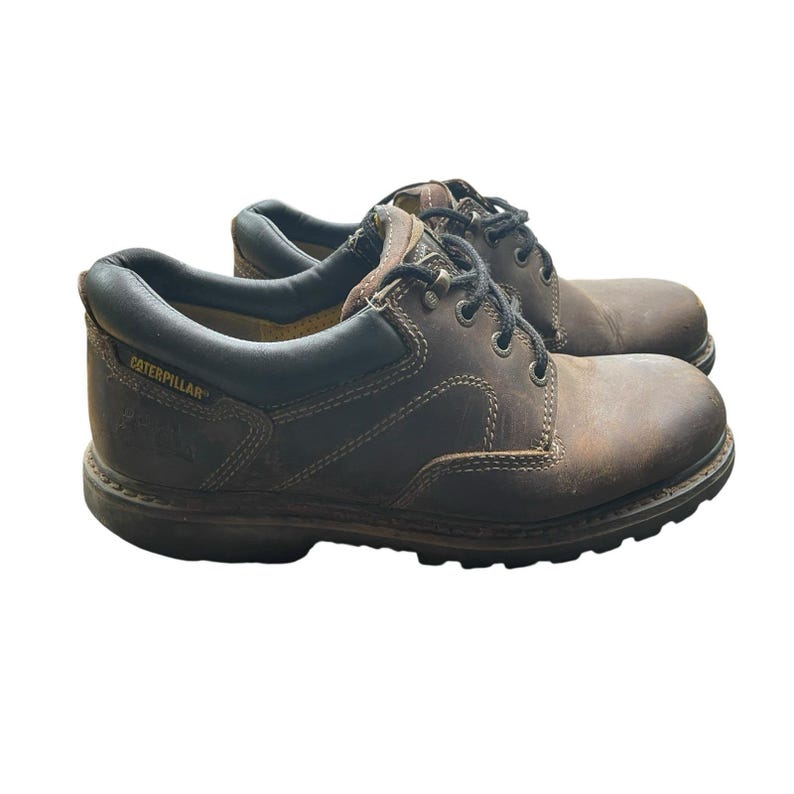 May include: Pair of brown leather work boots with black laces. The boots have a rounded toe and a thick, dark brown sole. The brand name "Caterpillar" is visible on the side of the boot.