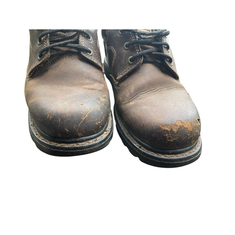 May include: Pair of worn brown leather work boots with black laces. The boots show signs of wear and tear, with scuffs and areas where the leather is peeling. The soles are black and thick, designed for durability.