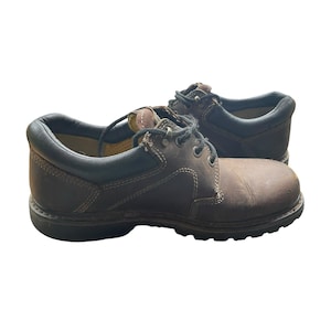 May include: A pair of brown leather work shoes with black trim. The shoes have laces and a sturdy sole, designed for durability and support. The leather has a worn appearance, suggesting regular use.