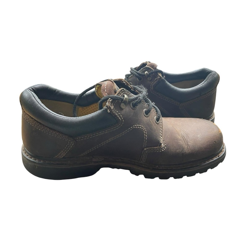 May include: A pair of brown leather work shoes with black trim. The shoes have laces and a sturdy sole, designed for durability and support. The leather has a worn appearance, suggesting regular use.