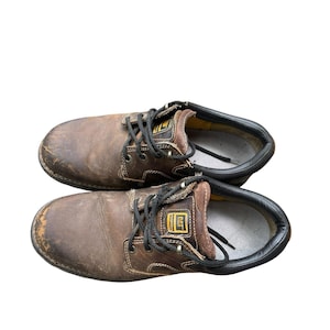 May include: Pair of brown leather work boots with black laces. The boots show signs of wear, with scuffs and creases on the leather. The tongue of each boot has a yellow and black logo. The soles are black and the interior is yellow.