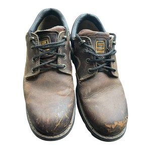 May include: Pair of brown leather work shoes with black laces. The shoes feature the CAT logo and show signs of wear. The soles are thick and durable, suitable for work. The shoes are designed for work environments.
