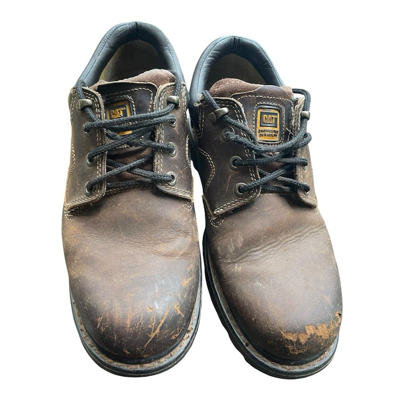May include: Pair of brown leather work shoes with black laces. The shoes feature the CAT logo and show signs of wear. The soles are thick and durable, suitable for work. The shoes are designed for work environments.